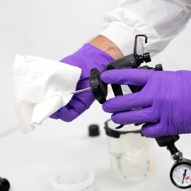 Hände mit violetten Handschuhen reinigen eine Lackierpistolennadel mit einem weißen Reinigungstuch