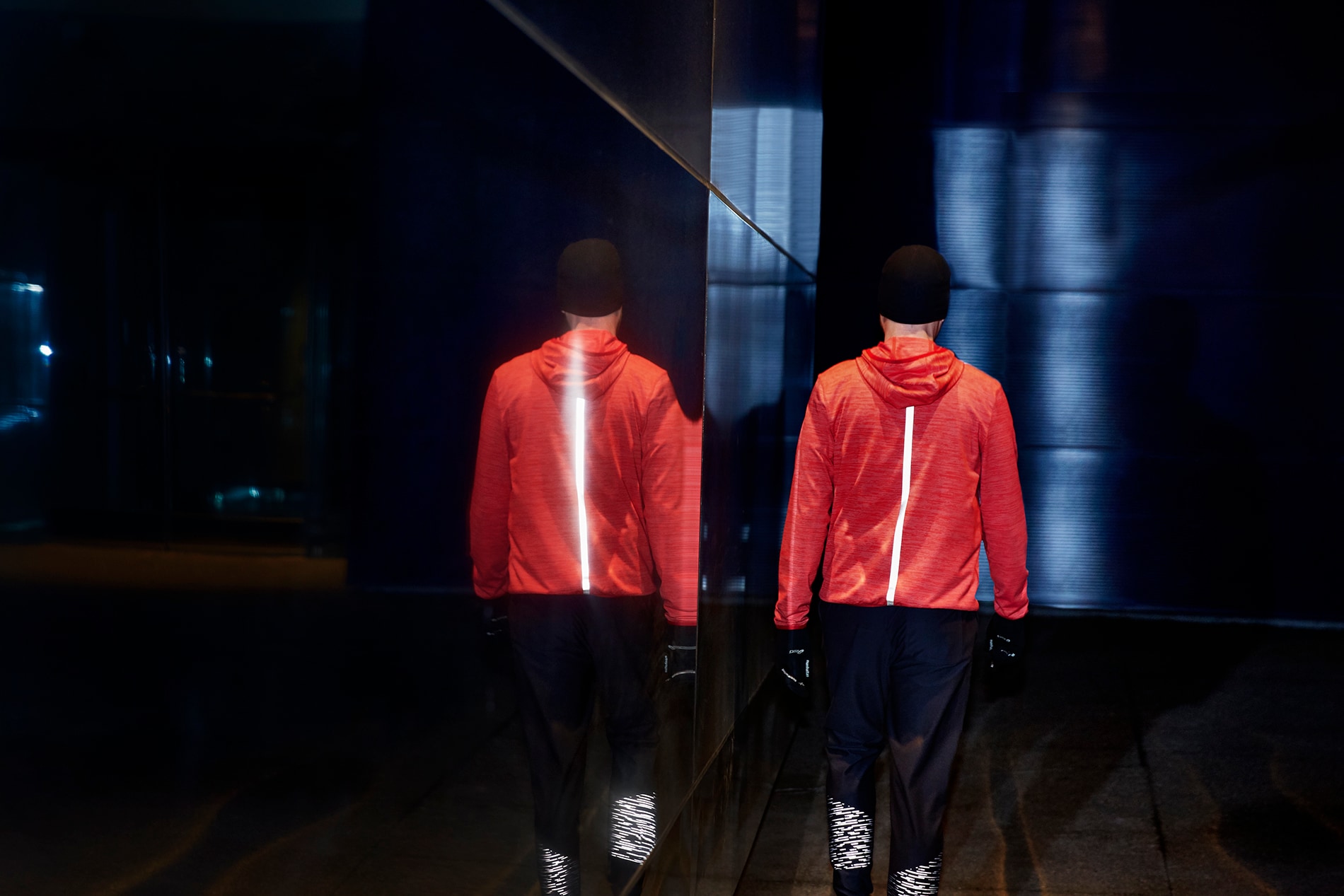 Beleuchteter Black Stretch Reflective Streifen auf dem Rücken einer Laufjacke eines Mannes, der an einer Spiegelwand bei Nacht vorbeigeht.