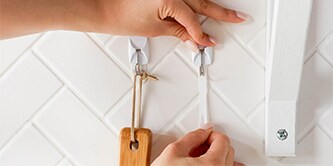Person, die zeigt, wie man mit Command™ Selbstklebende Haken und Strips organisiert.
