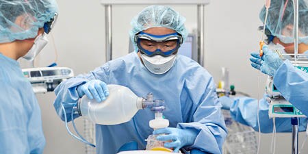 Das Bild zeigt drei medizinische Mitarbeiter, die Persönliche Schutzausrüstung tragen, während sie sich um einen Patienten kümmern.
