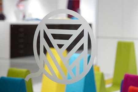 Klares Glasfenster mit Logo in Ätzglasoptik und bunten Formen dahinter.
