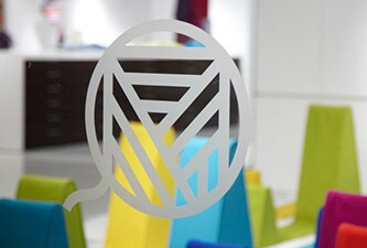 Klares Glasfenster mit Logo in Ätzglasoptik und bunten Formen dahinter.
