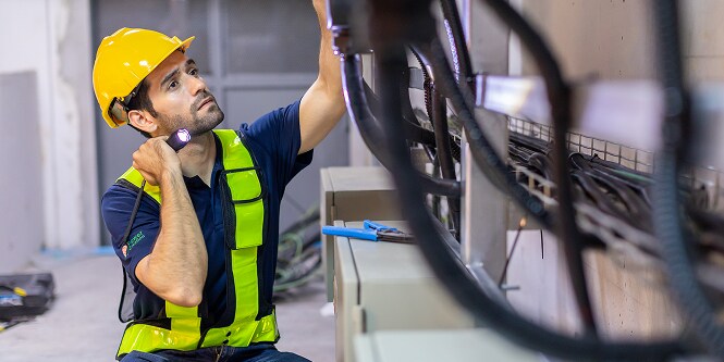 Arbeiter in Schutzkleidung, der mit einer Taschenlampe elektrische Anschlüsse überprüft
