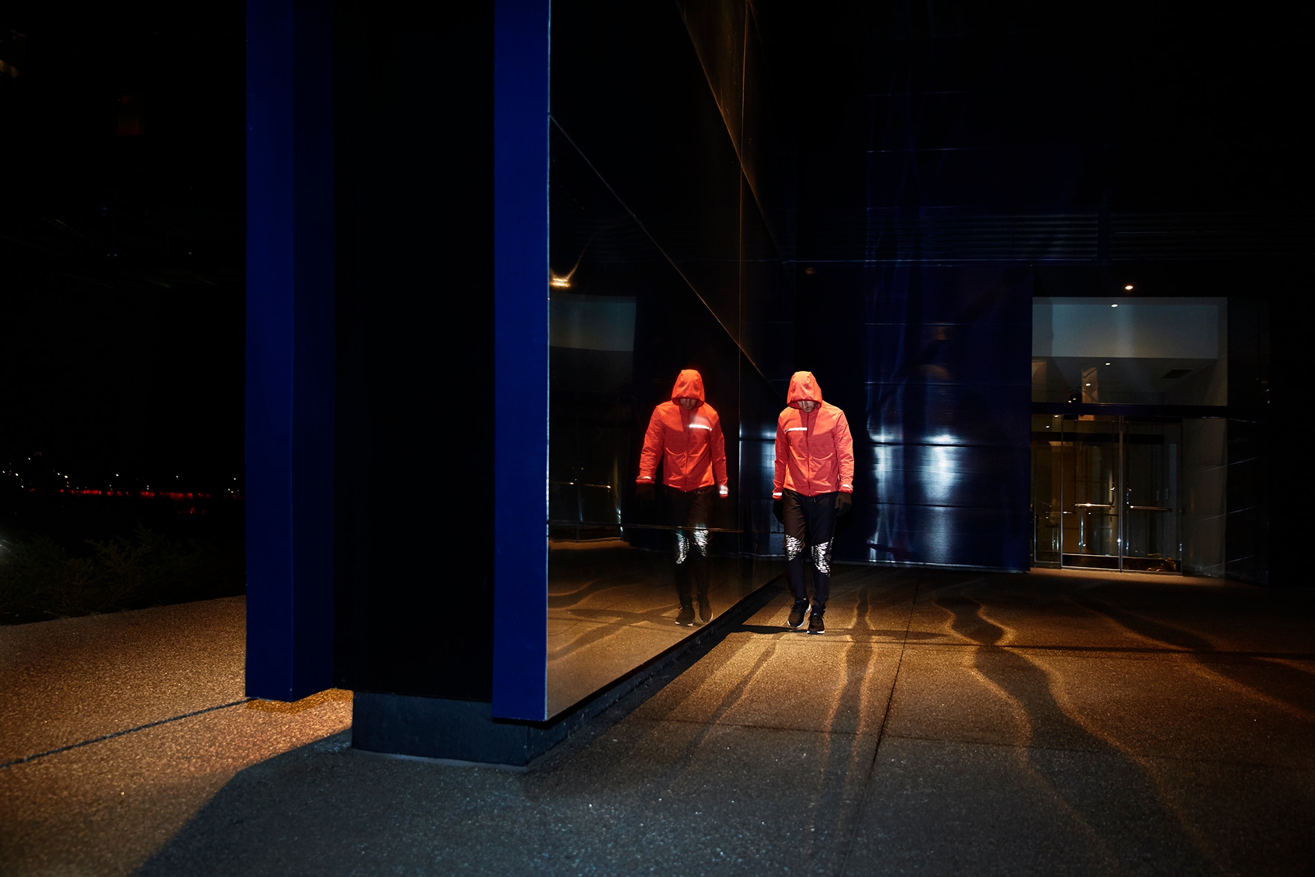 Beleuchtete Black Stretch Reflective Kniepaneele auf Lauftights eines Mannes, der an einer Spiegelwand bei Nacht vorbeigeht.
