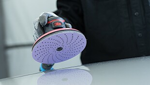 3M Exzenterschleifer mit perforierter Schleifscheibe, gegen eine glatte Oberfläche.
