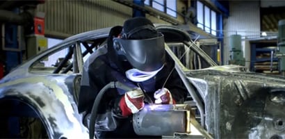 Bild mit einem Schweißer mit Schutzausrüstung, der an einer Metallstruktur in einer Fertigungsanlage einen Schweißbrenner anwendet
