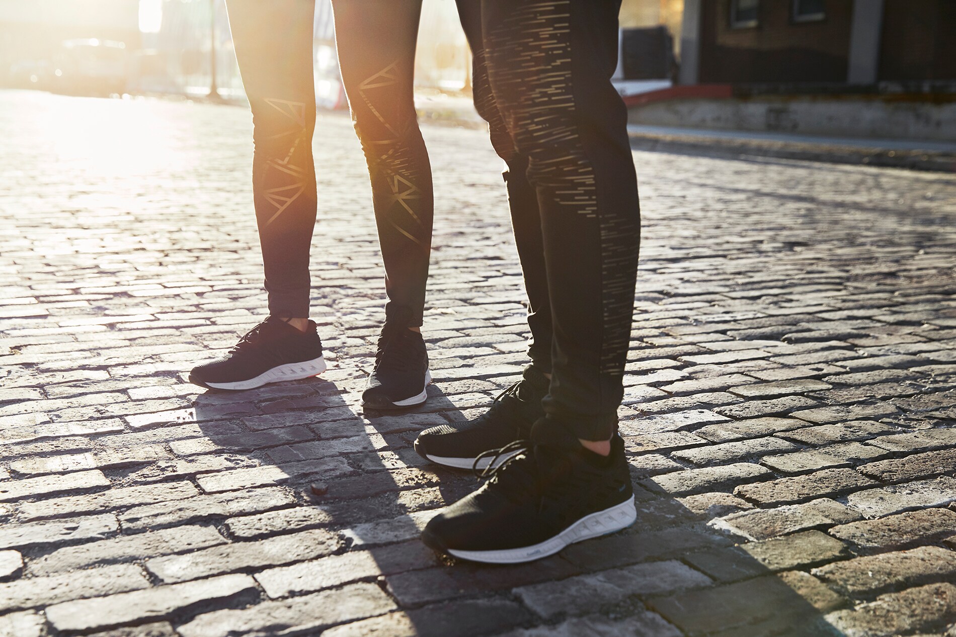 Mann und Frau stehen auf einer Kopfsteinpflasterstraße im Sonnenuntergang in Lauftights mit 3M Scotchlite Black Stretch Reflective.