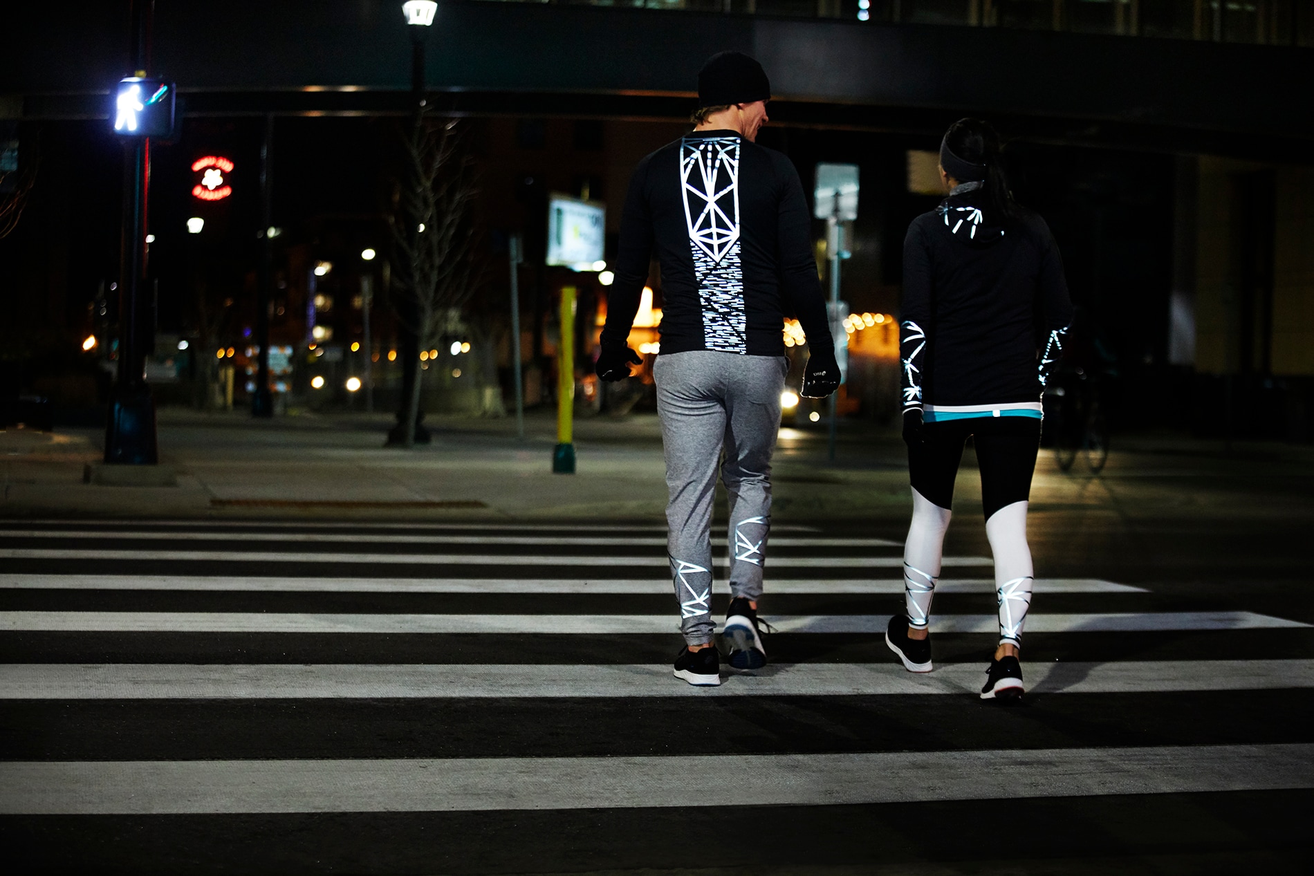 Mann und Frau laufen durch nächtliche Stadtlandschaft, Ausstattung beleuchtet von 3M Scotchlite Black Stretch Reflective.
