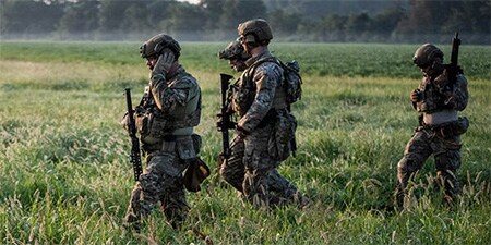 Das Bild zeigt vier  voll bewaffnete Soldaten im grasbewachsenen Gebiet mit modernen taktischen Headsets.
