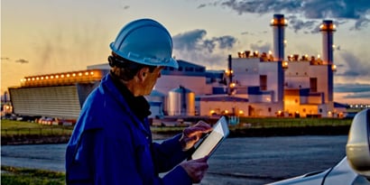 Mann mit Bauhelm auf einem iPad vor einer Industrieanlage 

